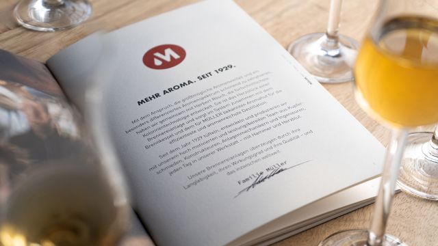 Aufgeschlagene Broschüre von Müller Brennereianlagen mit Firmenphilosophie und Signatur, umgeben von gefüllten Schnapsgläsern auf einem Holztisch.