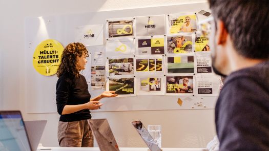 Eine Frau präsentiert während eines Recruiting-Workshops in einem Büro eine Wand mit Marketingentwürfen.