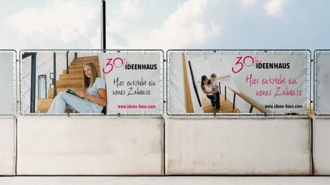 Bauzaunbanner von Ideenhaus mit Slogan „Hier entsteht ein neues Zuhause“ und Bildern von Menschen im modernen Treppenhaus.