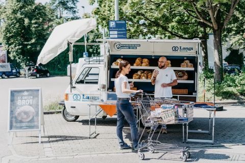 Verkaufsstand von Langs Backstube mit frischem Brot, Kunde mit Einkaufswagen im Gespräch mit Verkäufer.