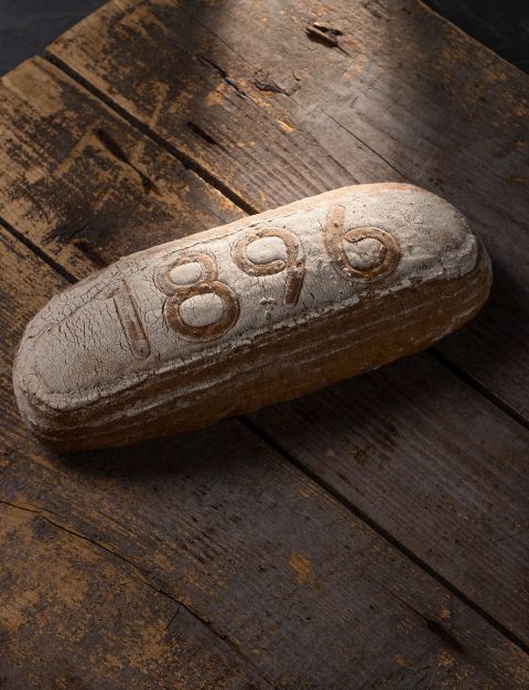 Ein großer Brotlaib, auf dem "1896" steht, liegt auf einem Holztisch.