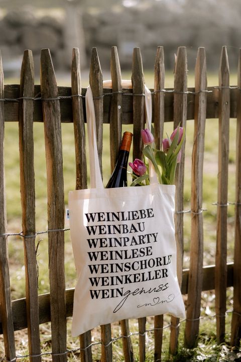 Tragetasche mit Text, die an einem Holzzaun hängt und eine Weinflasche und Tulpen enthält.