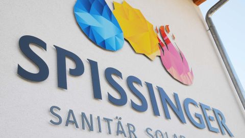 Logo der Firma Spissinger mit bunten Symbolen für Wasser, Sonne und Wärme auf heller Hauswand.