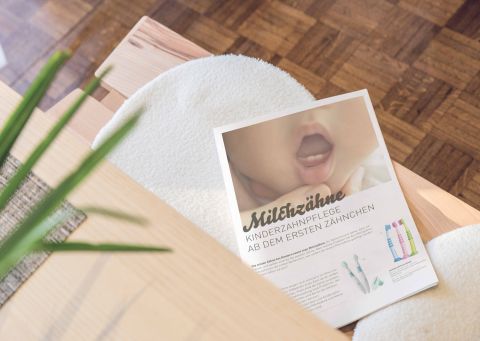 Eine Zeitschrift mit dem Titel „Milchzähne“ über Kinderzahnpflege liegt auf einem weißen Stuhl neben einem Holztisch.