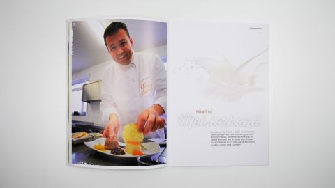 Offene Broschüre mit Bild eines lächelnden Kochs bei der Dessertanrichtung und Text „Produkte aus Meisterhand“.