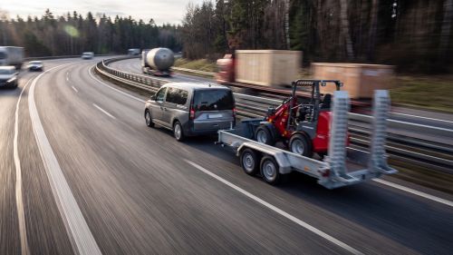 VW Caddy zieht einen Baumaschinenanhänger beladen mit einem roten Bagger auf der Autobahn.