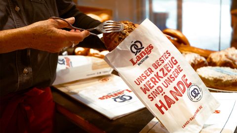 Eine Verkäuferin legt mit einer Zange ein frisch gebackenes Brot in eine Papiertüte von Langs Backstube mit der Aufschrift „Unser bestes Werkzeug sind unsere Hände“.