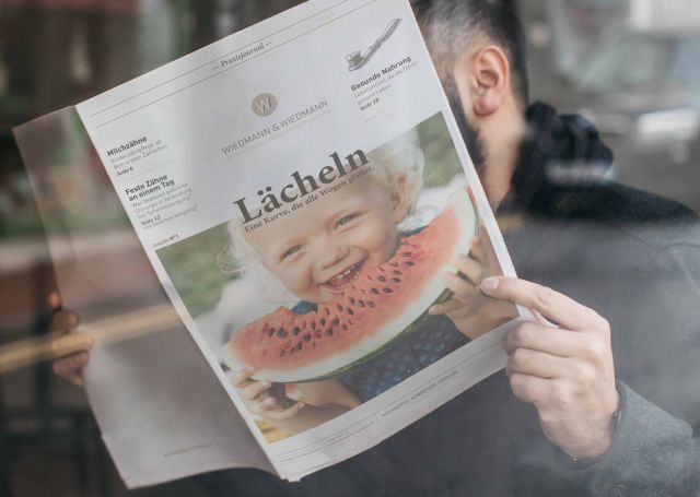 Ein Mann hält eine Zeitschrift mit dem Titel „Lächeln“, die ein lachendes Kind mit Wassermelone auf dem Cover zeigt.