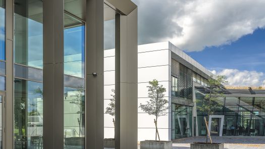 Modernes Bürogebäude mit Glasfassade, großen Metallstützen und bepflanzten Außenbereichen unter bewölktem Himmel.