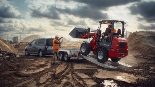 Ein roter Bagger von Weidemann wird über Rampen auf einen Hirth Baumaschinenanhänger gefahren.