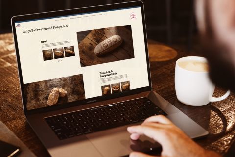 Ein Mann betrachtet die Website von Langs Backstube auf einem Laptop, mit Bildern von Brot und Gebäck, daneben eine Tasse Kaffee auf dem Holztisch.