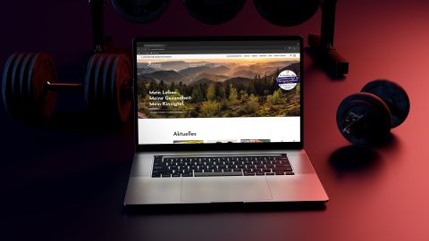 Ein Laptop mit geöffneter Gesundheitswebsite steht auf einem Tisch, im Hintergrund Hanteln auf dunklem roten Boden.