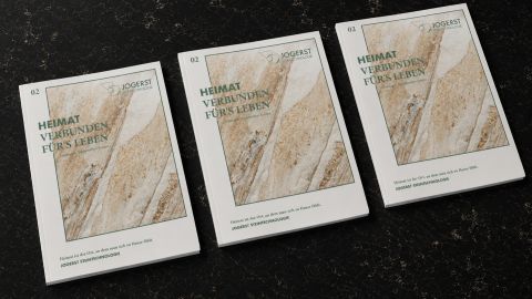Drei Ausgaben des Heimatkatalogs von Jogerst Steintechnologie zu Natursteinmaterialien aus Europa nebeneinander auf einem dunklen Stein-Hintergrund.