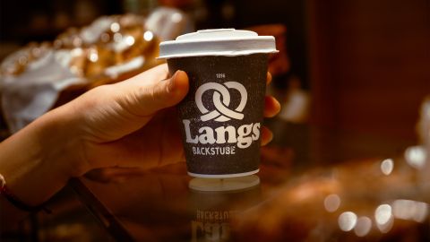 Eine Hand hält einen schwarzen Kaffeebecher mit weißem Deckel und dem Logo von Langs Backstube, im Hintergrund unscharfe Backwaren.