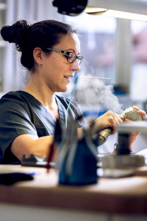 Eine Zahntechnikerin mit Brille arbeitet konzentriert im Labor mit einem Fräswerkzeug an einer Zahnprothese.
