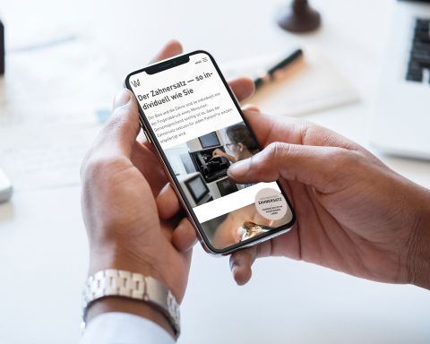 Eine Hand hält ein Smartphone, das eine Website über individuellen Zahnersatz anzeigt, mit Bildern aus einem Dentallabor.