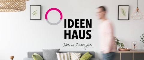 Ideenhaus-Logo mit dem Slogan „Ideen ein Zuhause geben“ vor stilvollem Wohnzimmerhintergrund mit Dekoration.