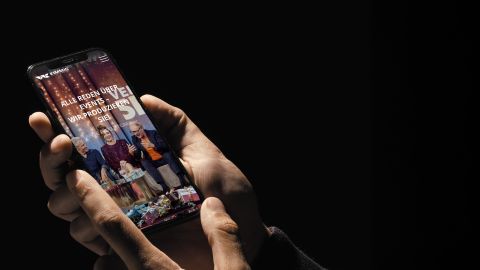Hand hält ein Smartphone mit einer Event-Produktionswebsite, die eine Talkshow mit drei Gästen und festlicher Dekoration zeigt.
