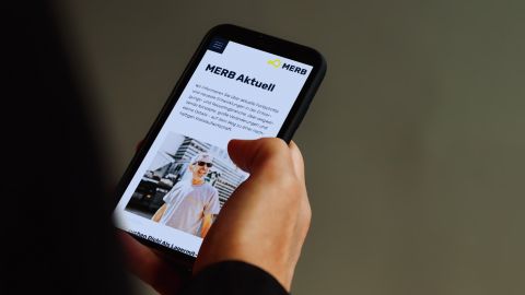 Hand hält Smartphone mit geöffneter MERB-Webseite, die aktuelle Informationen und ein Bild eines Mitarbeiters zeigt.