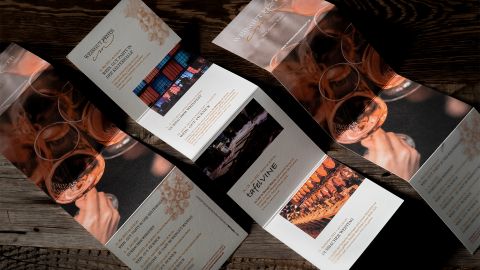 Aufgeklappte Event-Broschüren mit edlem Design, Weinverkostungsbildern und Texten, auf rustikalem Holztisch arrangiert.