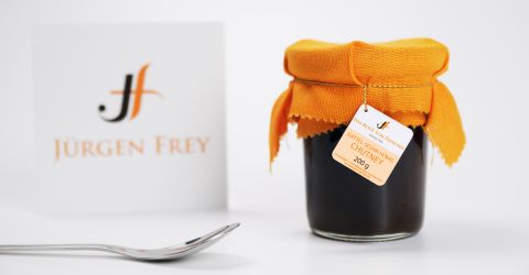 Dattel-Sesam-Honig-Chutney von Jürgen Frey mit orangem Stoffdeckel und Etikett, daneben Markenaufsteller und Löffel.