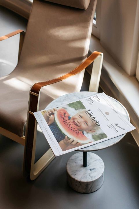 Eine Zeitschrift mit dem Titel „Lächeln“ und einem lachenden Kind mit Wassermelone liegt auf einem runden Marmortisch neben einem beigen Sessel.