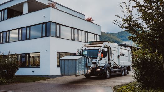 Ein Müllwagen von Weber hebt einen großen Metallcontainer vor einem modernen Bürogebäude mit Grünflächen.