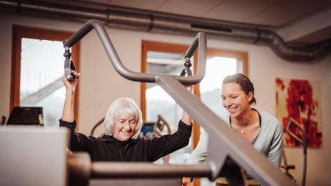 Ältere Frau trainiert an einem Fitnessgerät, unterstützt von einer lächelnden Trainerin in einem hellen Fitnessstudio.