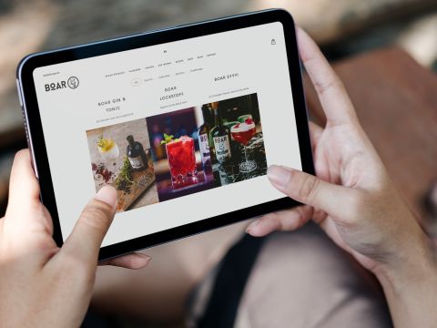Tablet in Händen zeigt die BOAR Gin Website mit Drinks wie Gin Tonic, roten Cocktails und BOAR Gin Flaschen.