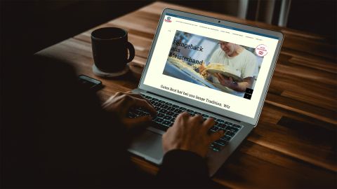 Eine Person nutzt einen Laptop, um die Website von Langs Backstube zu besuchen, mit dem Slogan „Feingebäck aus Meisterhand“. Eine Kaffeetasse steht daneben.