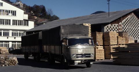 Historische Aufnahme von LKW im Palettenwerk Mercedes-LKW mit Plane vor einem Sägewerk, umgeben von gestapeltem Holz und Palettenmaterial Mitte des 20. Jahrhunderts.