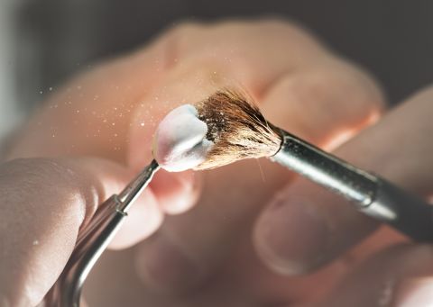 Nahaufnahme einer Zahntechnikerhand, die mit einem feinen Pinsel eine keramische Zahnkrone sorgfältig bearbeitet.