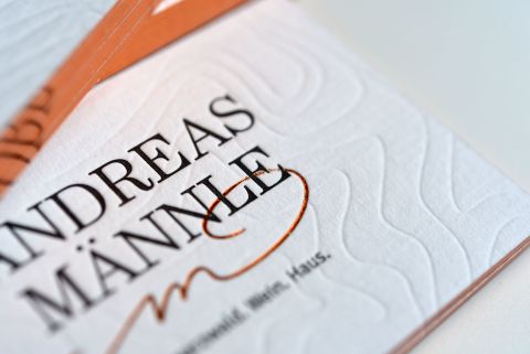 Nahaufnahme der Prägung auf einer weißen Visitenkarte mit der Aufschrift "Andreas Männle", einem kupferfarbenem "m" und Rand.