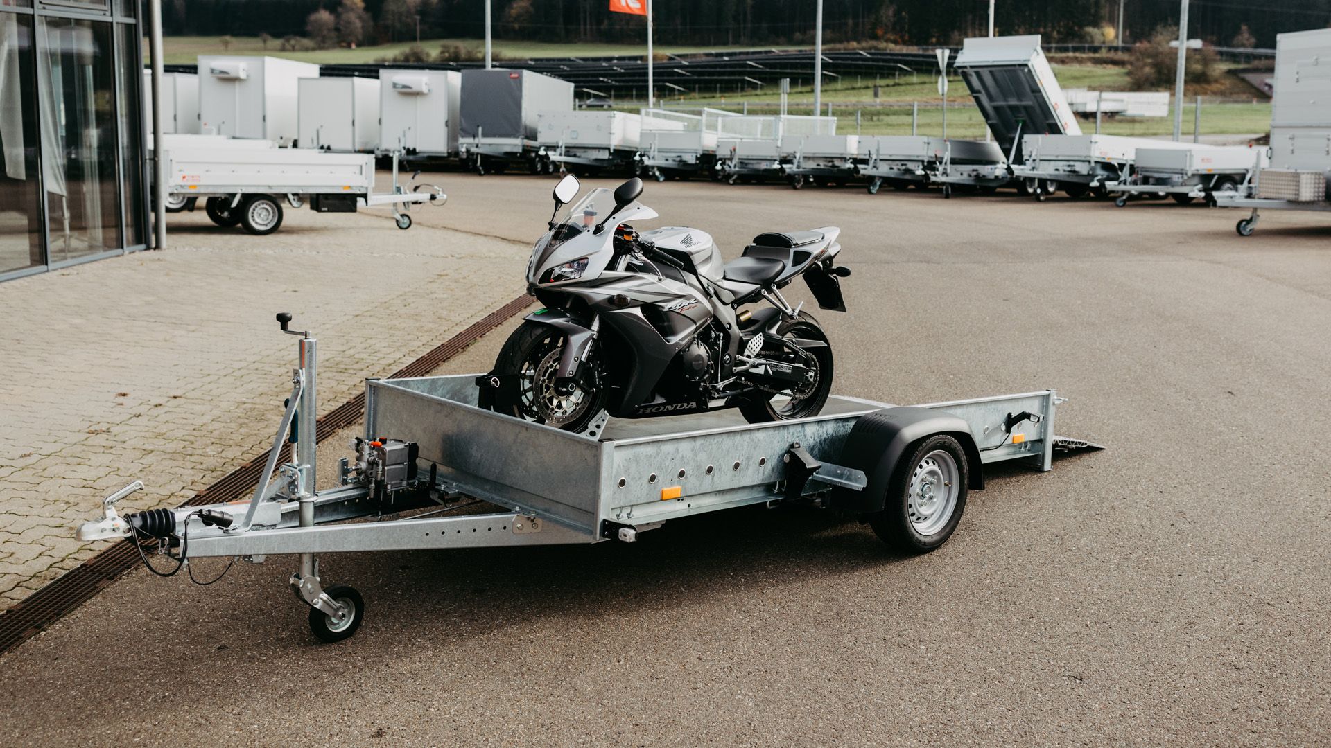 Ein silbernes Motorrad steht auf einem Absenk-Anhänger mit Auffahrrampen, geparkt auf dem Ausstellungsgelände vor dem Hirth-Firmengebäude.