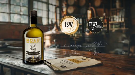 Eine Flasche BOAR Gin steht auf einem Holztisch in einem abgedunkelten Raum, daneben die Logos von der ISW-Auszeichnung "Gin des Jahres" im Hintergrund verschwommen ein Brennkessel und Holzfässer.