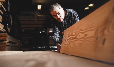 Mann prüft sorgfältig eine massive Holzbohle mit markanter Maserung in einer Werkhalle