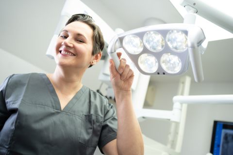 Eine lächelnde Zahnärztin in grauer Arbeitskleidung justiert die OP-Leuchte in einer modernen Zahnarztpraxis.