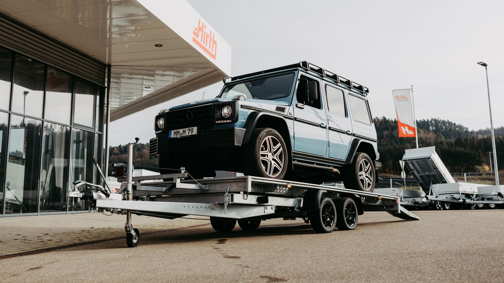 Ein hellblauer Geländewagen steht auf einem Autotransport-Anhänger mit Auffahrrampen, geparkt auf dem Ausstellungsgelände vor dem Hirth-Firmengebäude.