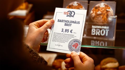 Eine Person hält ein Preisschild für „Bartholomäus Brot“ mit einem Preis von 3,95 €, während im Hintergrund Brote in der Bäckerei ausgestellt sind.