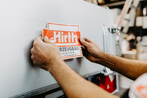 Nahaufnahme, wie ein rot-weißer Firmenaufkleber mit Logo und Kontaktdaten von HIRTH auf die Bordwand eines silbernen Pkw-Anhängers geklebt wird.