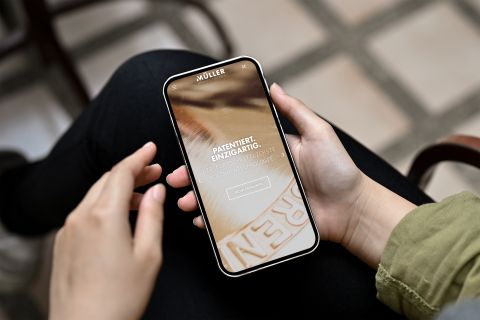 Müller Brennereianlagen-Website auf Smartphone Person hält ein Smartphone mit der Website von Müller Brennereianlagen, die ein Kupferelement und Werbetext zeigt.