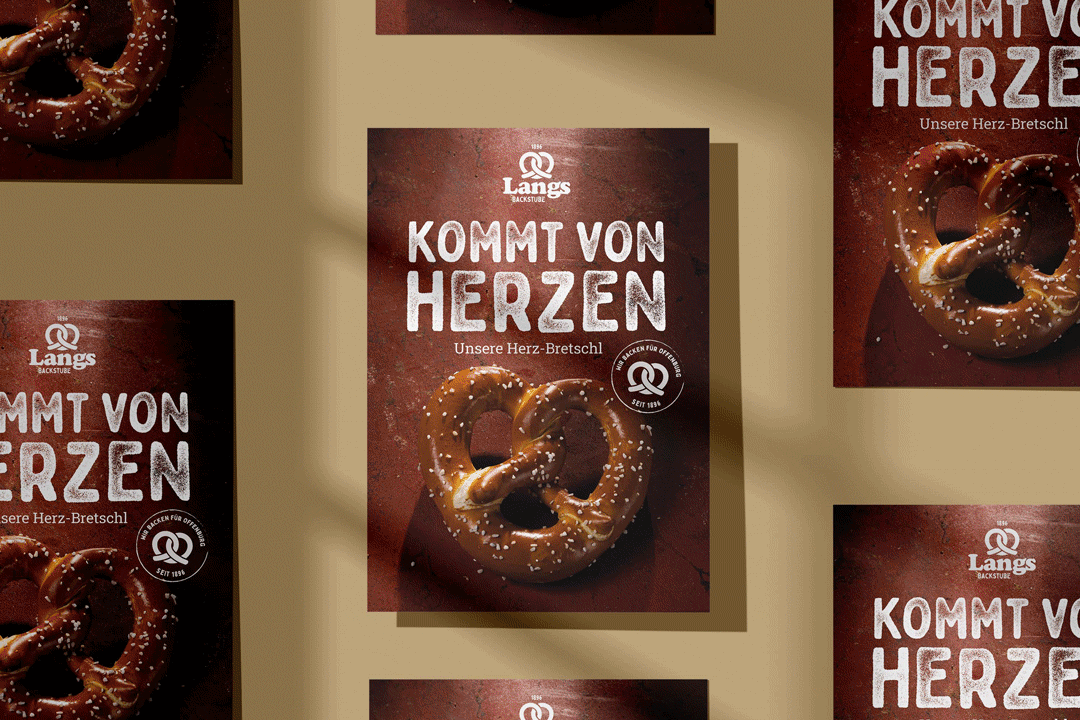Mehrere Werbeplakate von Langs Backstube mit einer herzförmigen Brezel und dem Slogan „Kommt von Herzen“ auf braunem Hintergrund.