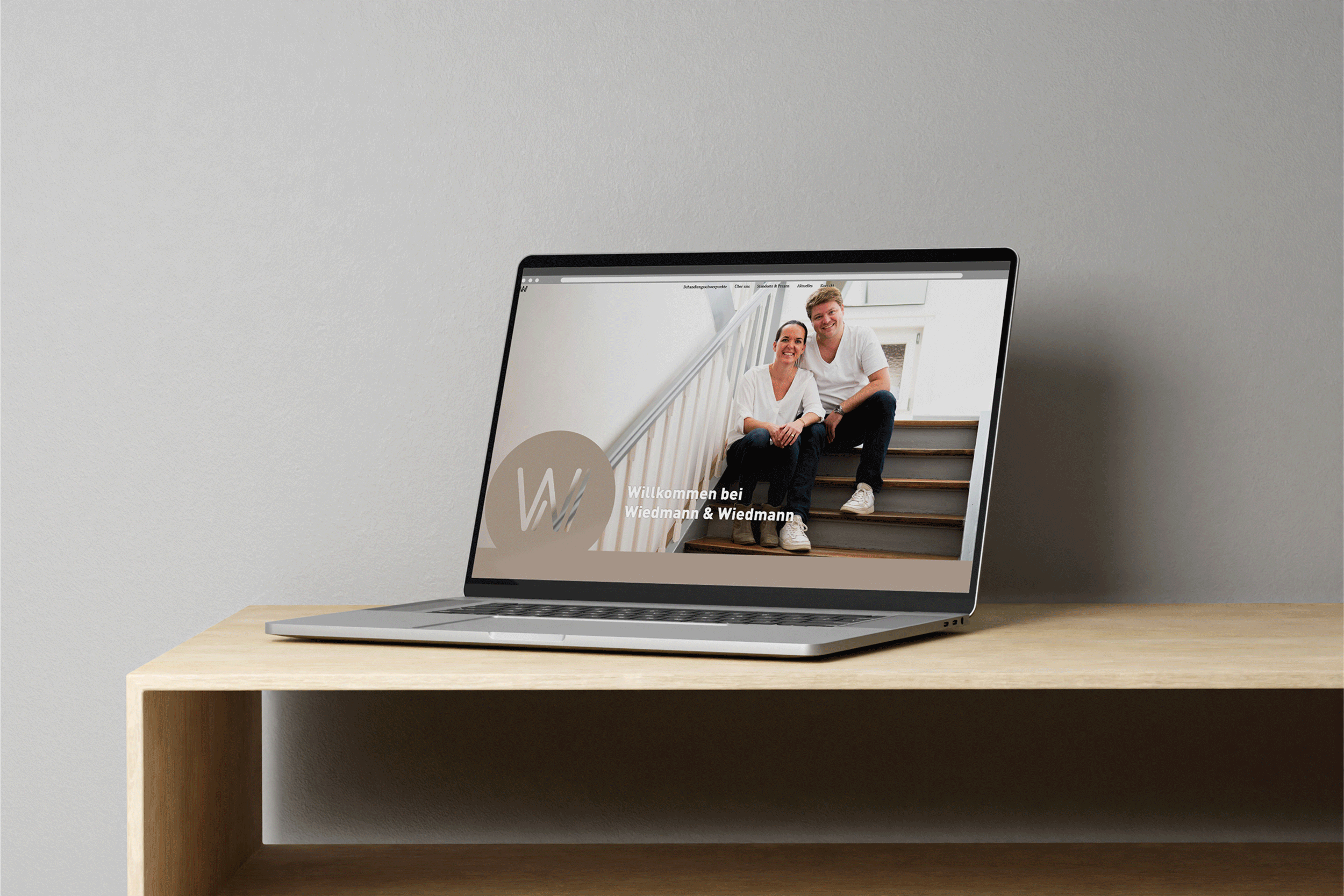Ein Laptop auf einem Holztisch zeigt die Website von Wiedmann & Wiedmann mit einem Begrüßungsbild von zwei Personen auf einer Treppe.
